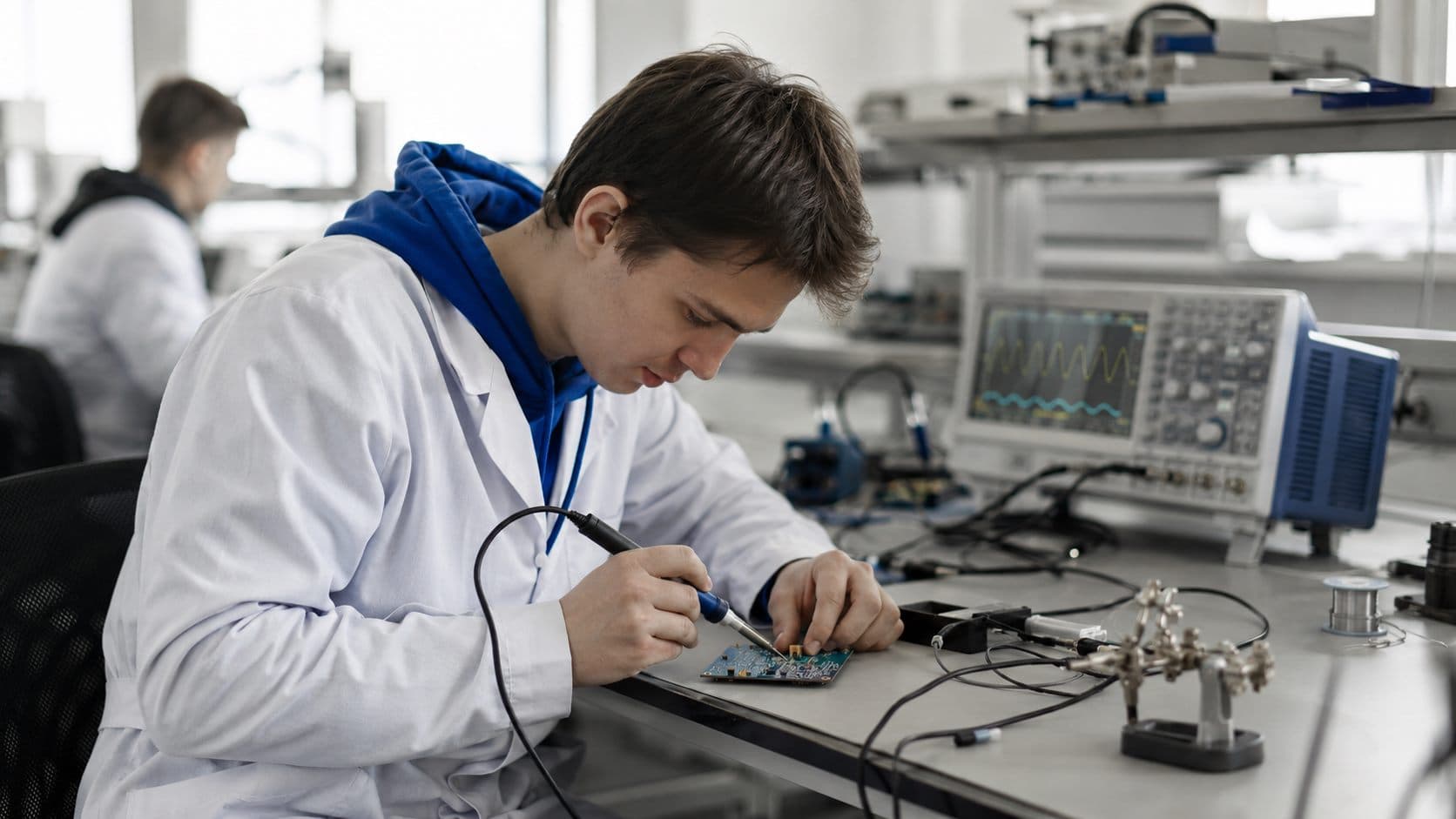 Студент паяет плату за электронным верстаком с осциллографом